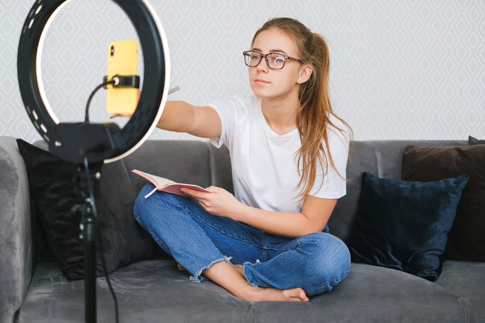 Woman vlogging with smartphone and ring light while holding a book on a sofa.