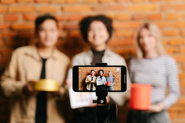 A diverse team of vloggers recording a video with a smartphone against a brick wall backdrop. Influencer marketing per i brand italiani